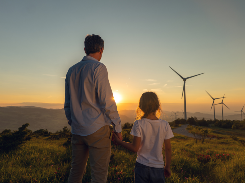 A father and daughter hold hands and look out over a field with wind turbines