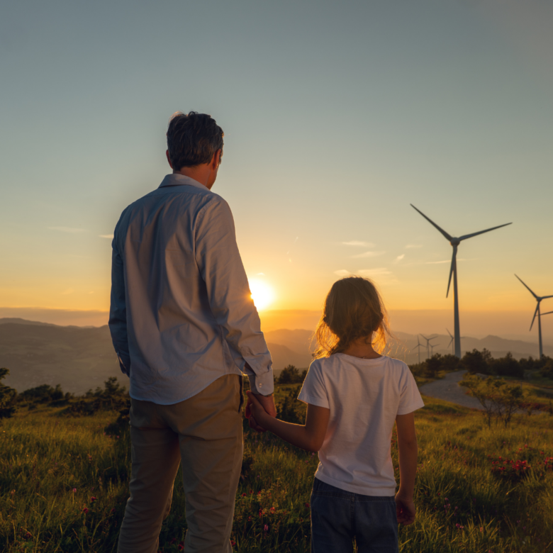 A father and daughter hold hands and look out over a field with wind turbines