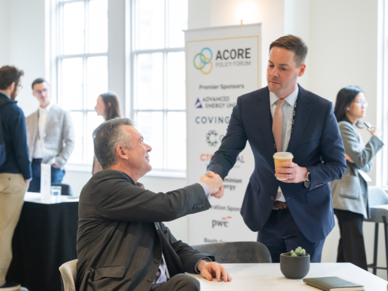 Attendees shake hands during a networking break with a sponsor booth in the background.