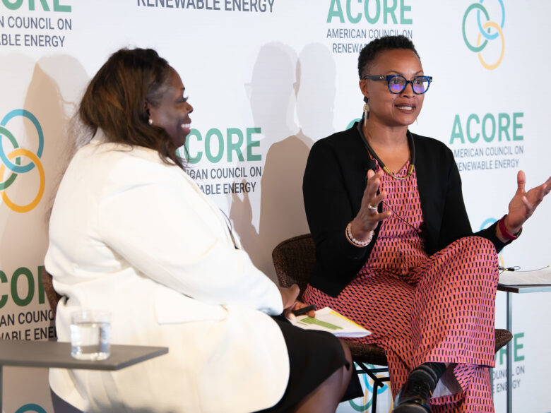 Two women sitting on chairs talking to each other in front of a backdrop with the ACORE logo.