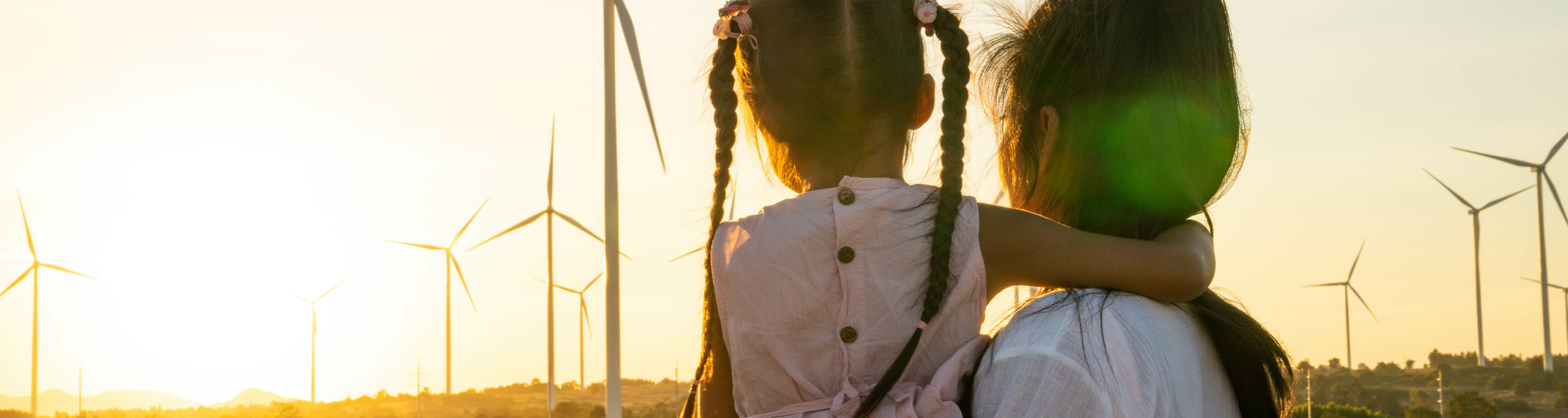 A woman holding a little girl with two pigtail braids in front of wind turbines at sunset.