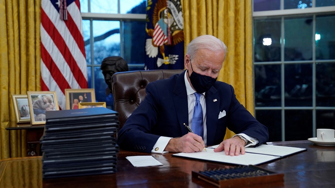 President Biden wearing a mask and sitting at the desk in the Oval Office, signing Executive Orders