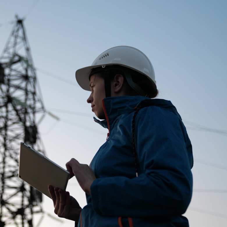 A woman in a hard hat is holding a tablet in front of power lines.