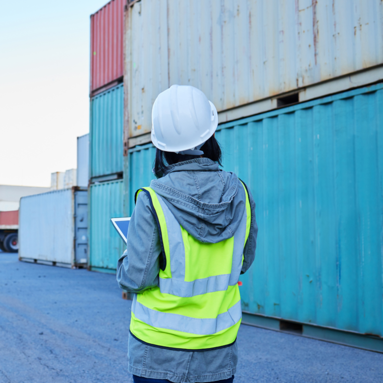 A construction worker holding a tablet in front of shipping containers.