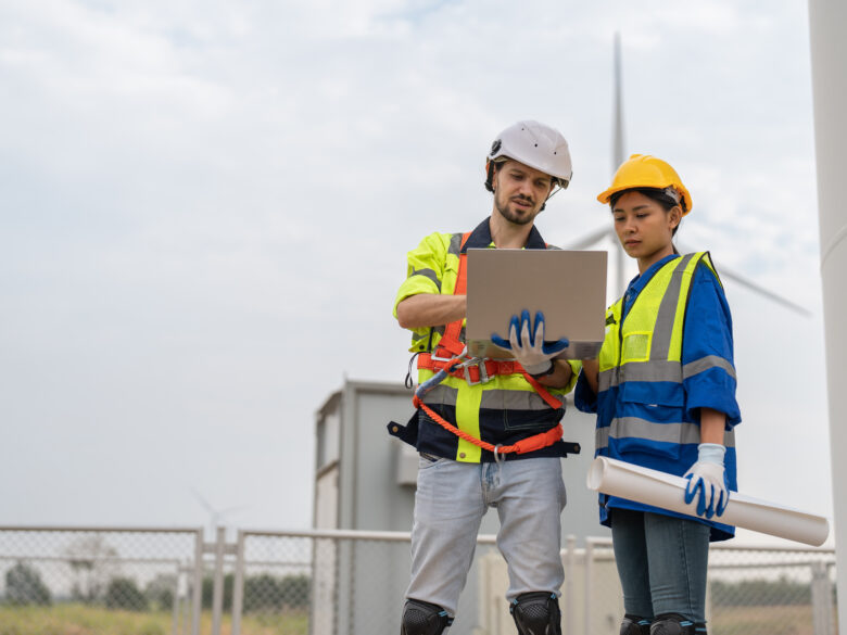 Male and female engineer in uniform with helmet safety using laptop discussing inspection and maintenance of wind turbine in wind farm to generate electrical energy, Renewable power energy