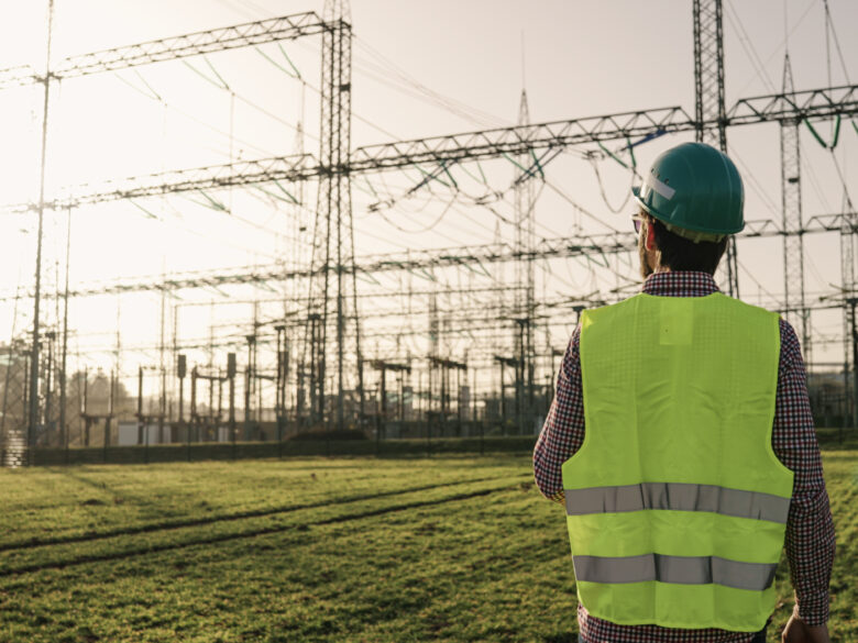 Electrical engineer wearing a helmet and safety vest working with tablet near high voltage electrical lines power station during sunset