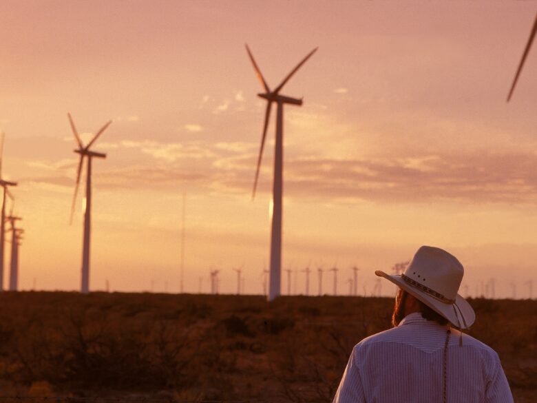 View of a man from behind wearing a cowboy hat and looking onto a field of wind turbines at sunset