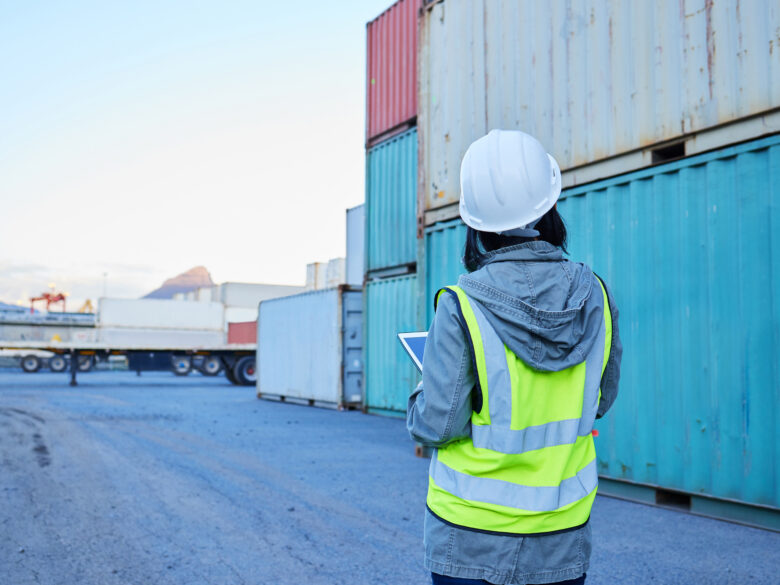 View of a woman from behind wearing a hard hat and a safety vest, standing in front of shipping containers