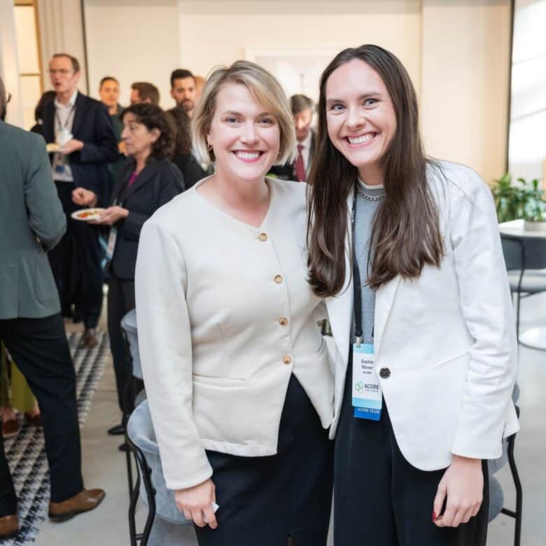 Two attendees smiling for a photo at the ACORE Grid Forum with others enjoying cocktail hour in the background.
