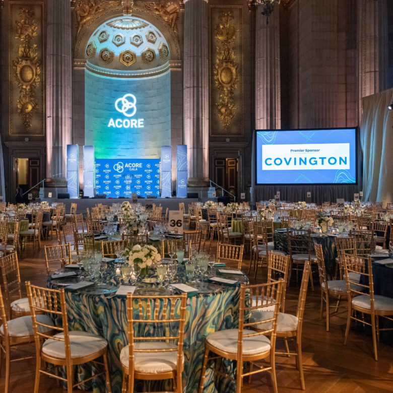 Round tables with blue-green patterned linens, floral centerpieces, and gold chairs fill the room. At the front, a lit stage displays ACORE branding, while a large screen highlights Covington as the premier sponsor.