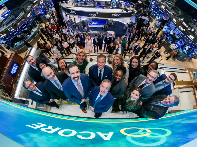 A wide, overhead group photo on the New York Stock Exchange trading floor shows ACORE board members, staff, and guests gathered beneath the NYSE and CNBC broadcast booths. ACORE branding is displayed on large digital screens throughout the trading floor as attendees look up and smile at the camera, commemorating ACORE's 25th anniversary and Opening Bell ceremony.