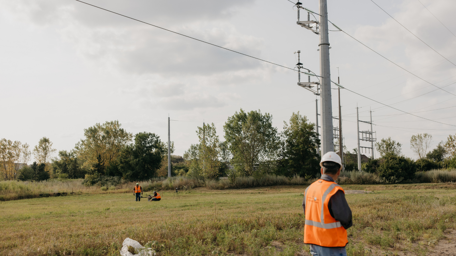 Workers in safety vests and hard hats conduct a dynamic line rating demonstration beneath high-voltage transmission lines in an open field.