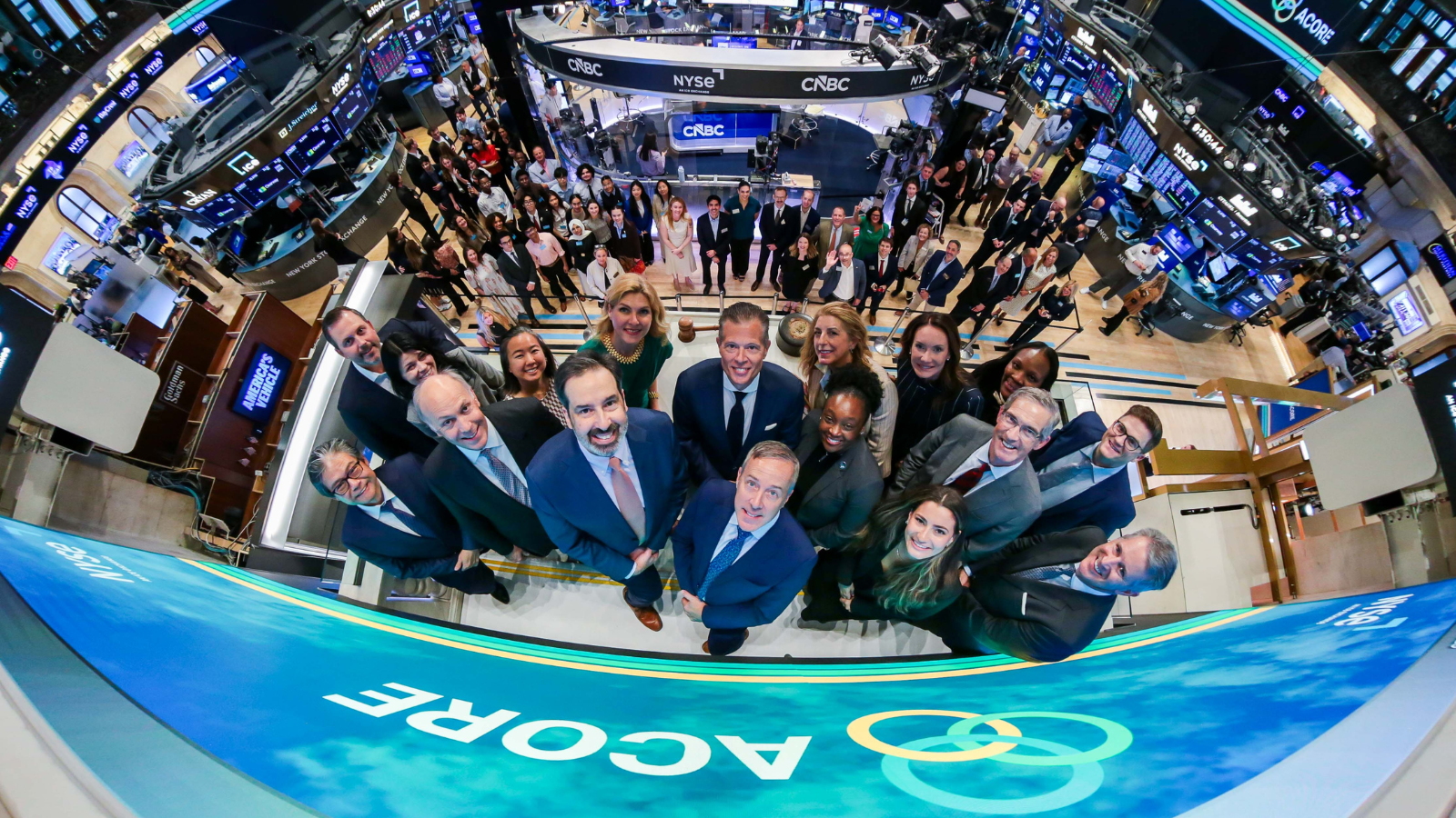A wide, overhead group photo on the New York Stock Exchange trading floor shows ACORE board members, staff, and guests gathered beneath the NYSE and CNBC broadcast booths. ACORE branding is displayed on large digital screens throughout the trading floor as attendees look up and smile at the camera, commemorating ACORE's 25th anniversary and Opening Bell ceremony.