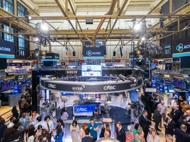 A wide view of the New York Stock Exchange trading floor. Multiple digital screens display the ACORE logo above the trading floor, while the circular CNBC and NYSE broadcast desk sits at the center.