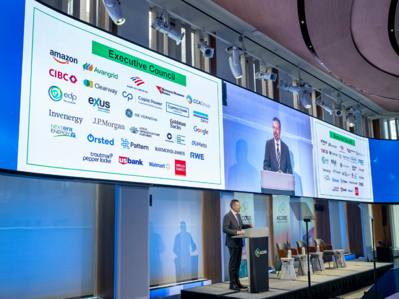 Speaker presenting on stage at the ACORE Finance Forum, with Executive Council member logos displayed on large screens.