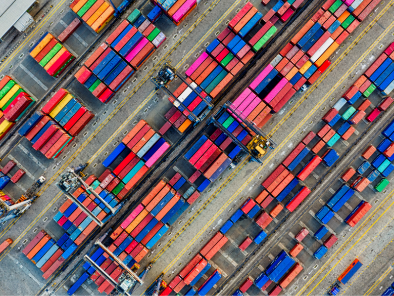 Aerial view of a shipping terminal with rows of colorful cargo containers, container-handling cranes, and marked transport lanes.