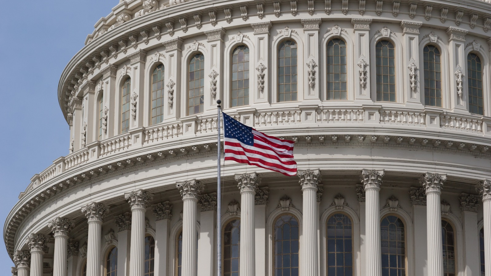 Close-up of the U.S. Capitol Building in Washington, D.C., featuring ornate white columns, arched windows, and an American flag waving in the foreground.