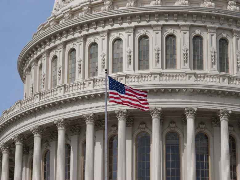 Close-up of the U.S. Capitol Building in Washington, D.C., featuring ornate white columns, arched windows, and an American flag waving in the foreground.