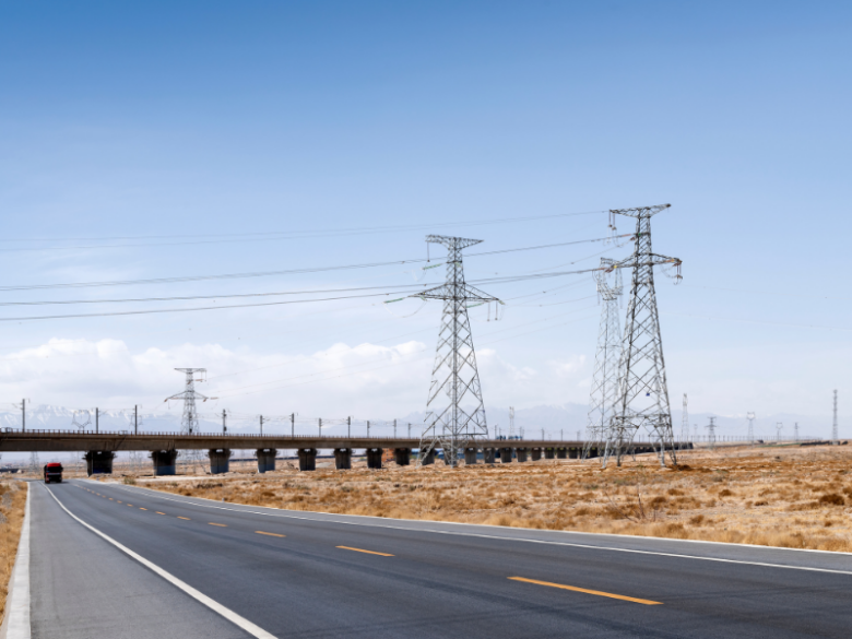 High-voltage electric transmission towers alongside a highway, with an elevated transportation corridor in the background.